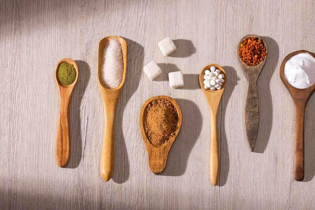 O que seria um bom adoçante? Por Paty Ayres Imagem de colheres de madeira contendo diversos tipos de açúcares e adoçantes - Por Paty Ayres